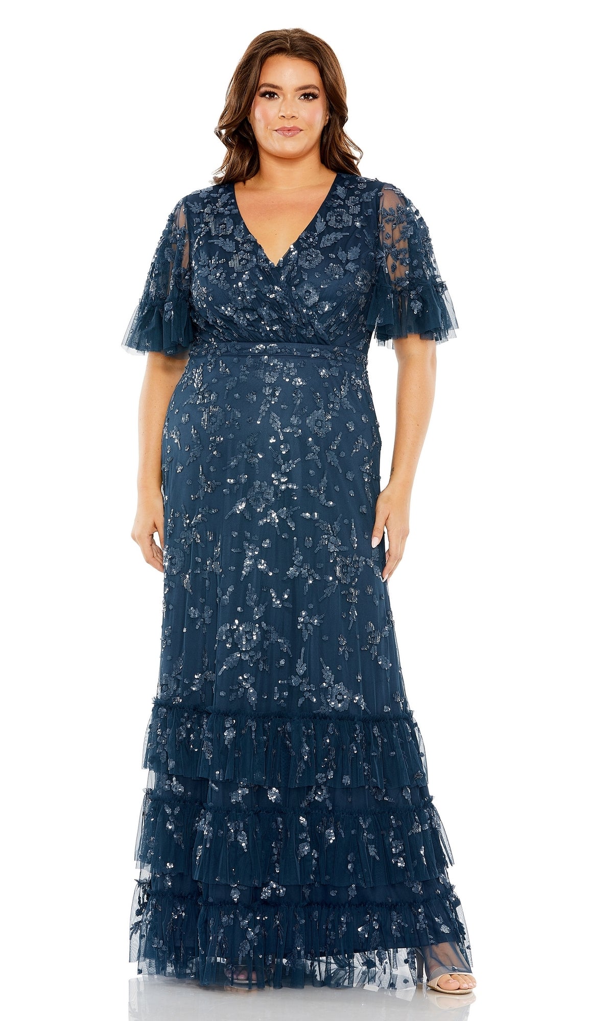 A woman stands against a white background wearing the Long Plus-Size Formal Dress 9270 by Mac Duggal: a floor-length, navy blue A-line dress with floral embroidery, layered ruffle details, and a v-neckline.